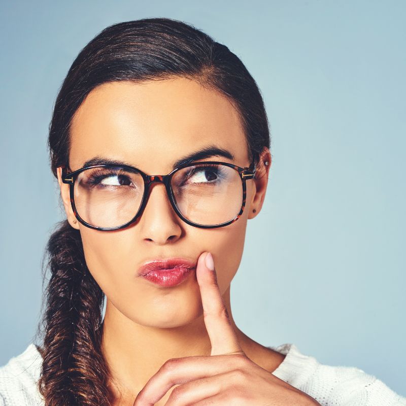 A woman with glasses on with her finger to her chin deep in thought.